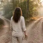 Une femme vue de dos à une fourche de sentier forestier dans la lumière d'un matin d'automne, carnet à la main